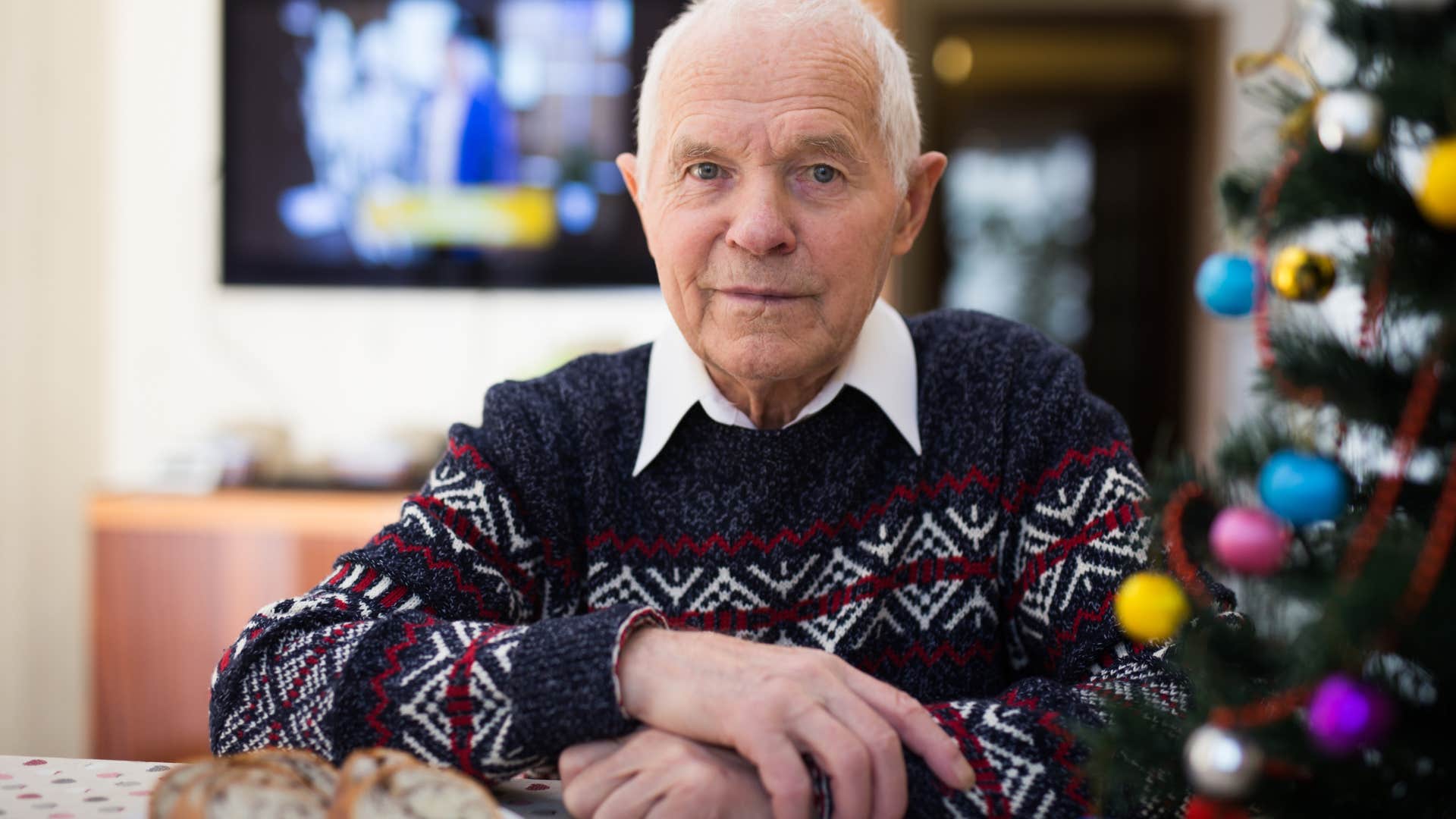 Older man sitting next to a Christmas tree looking upset.