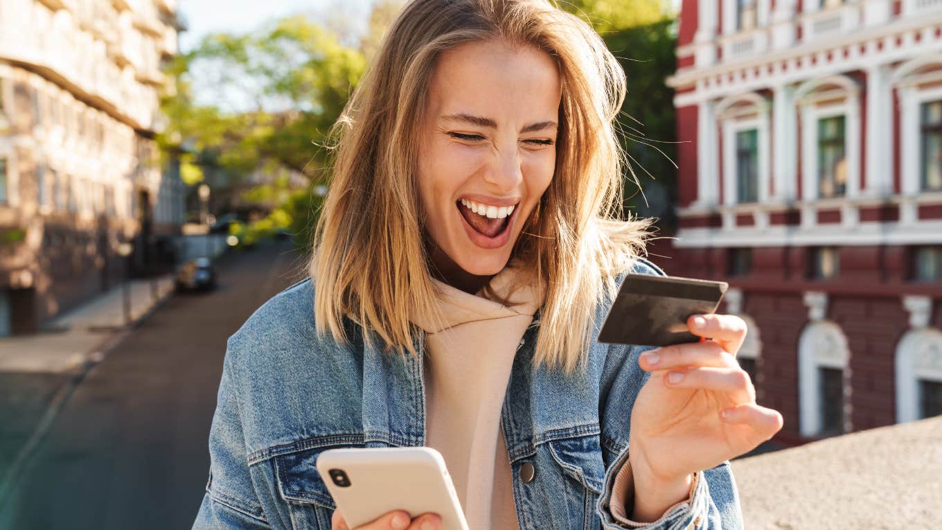 Beautiful cheerful young blonde woman wearing denim jacket walking in the city, using mobile phone, showing plastic credit card