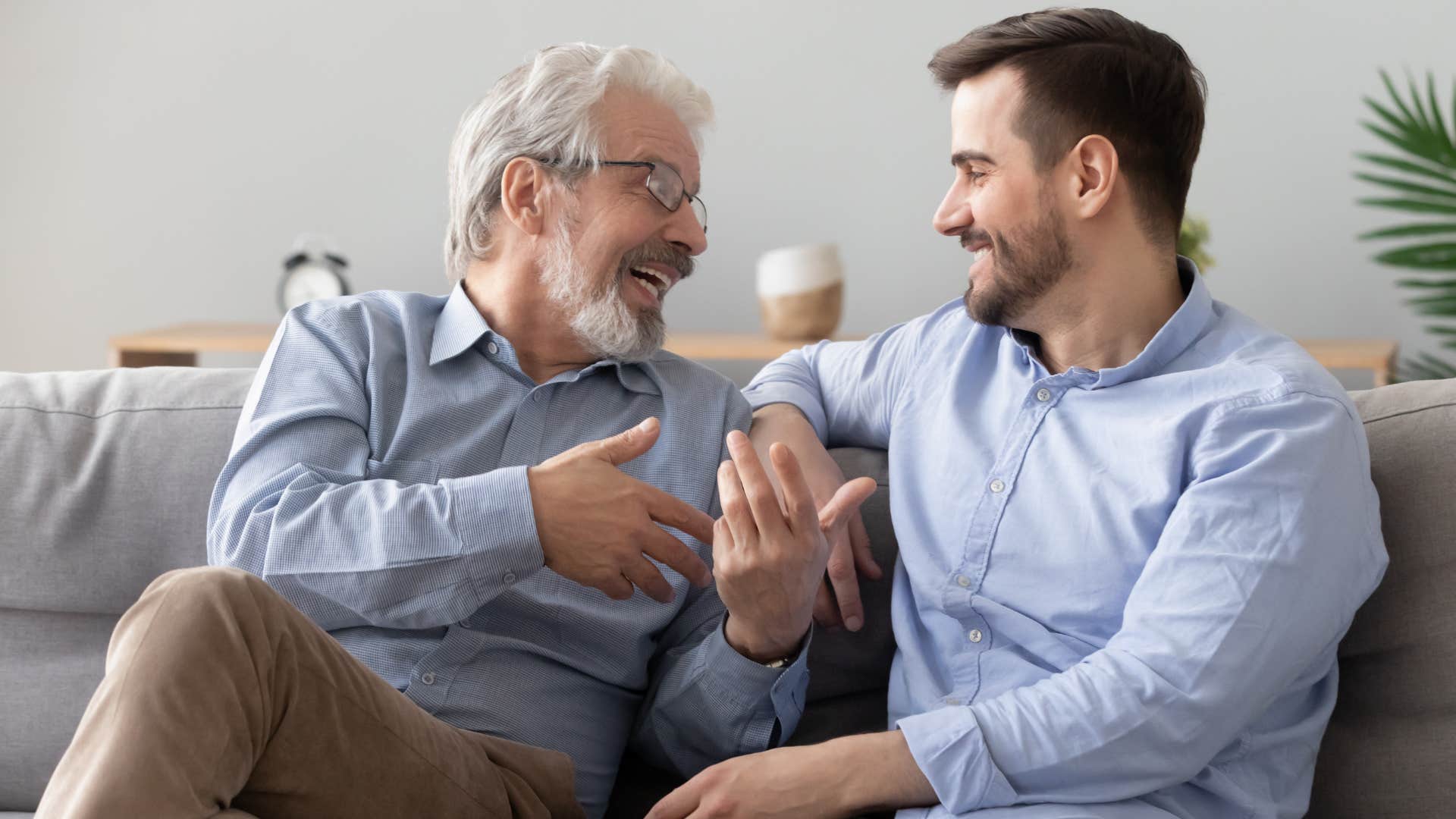 Adult son smiling and talking to his dad on the couch.