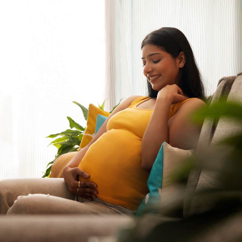 Smiling pregnant woman sitting on sofa at home with holding her abdomen