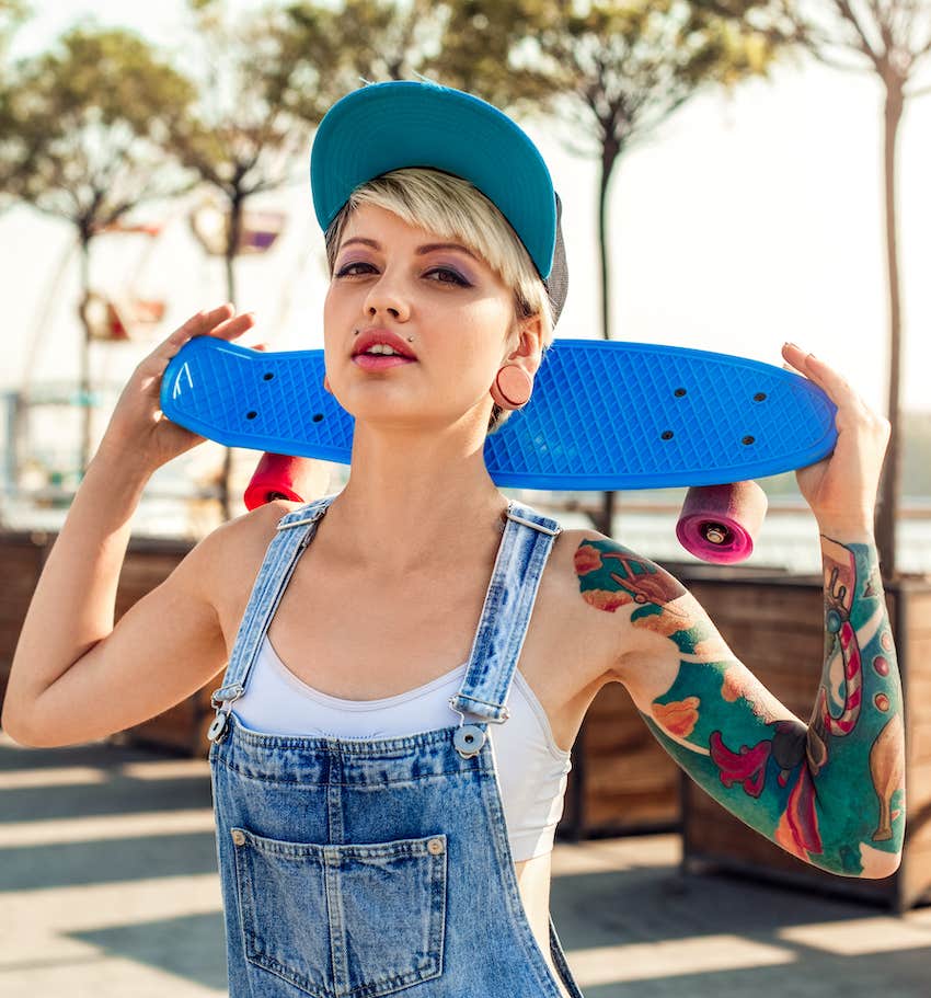 Woman in overalls holds skateboard behind her neck