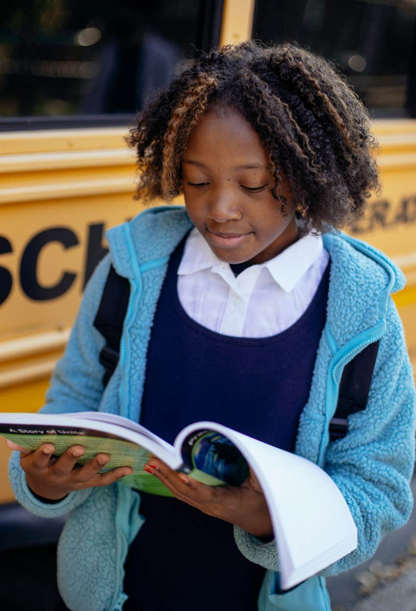 Young Black woman standing in front of school bus reading book