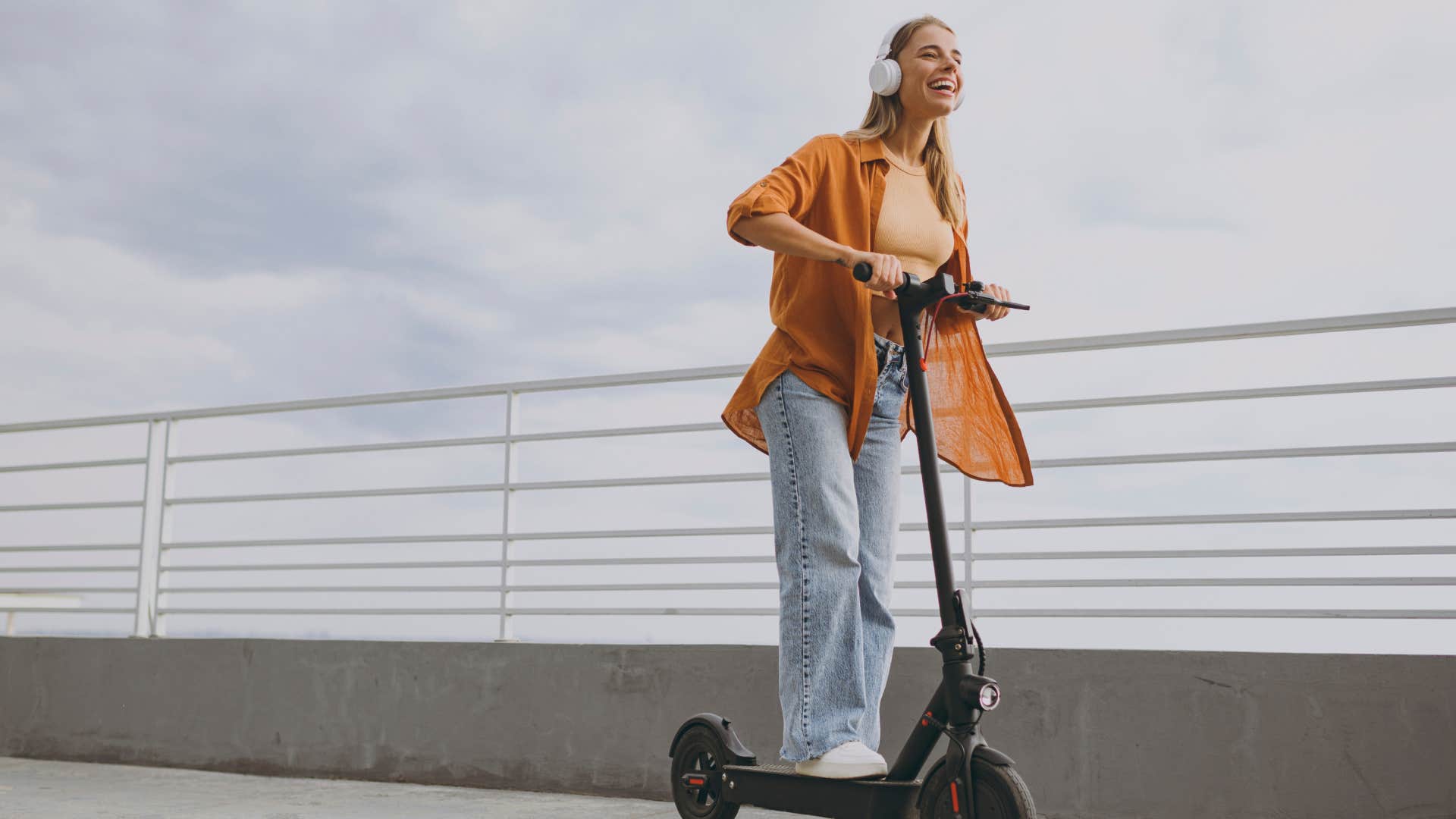 Full body young woman wears orange shirt casual clothes riding e-scooter transport listen music in headphones