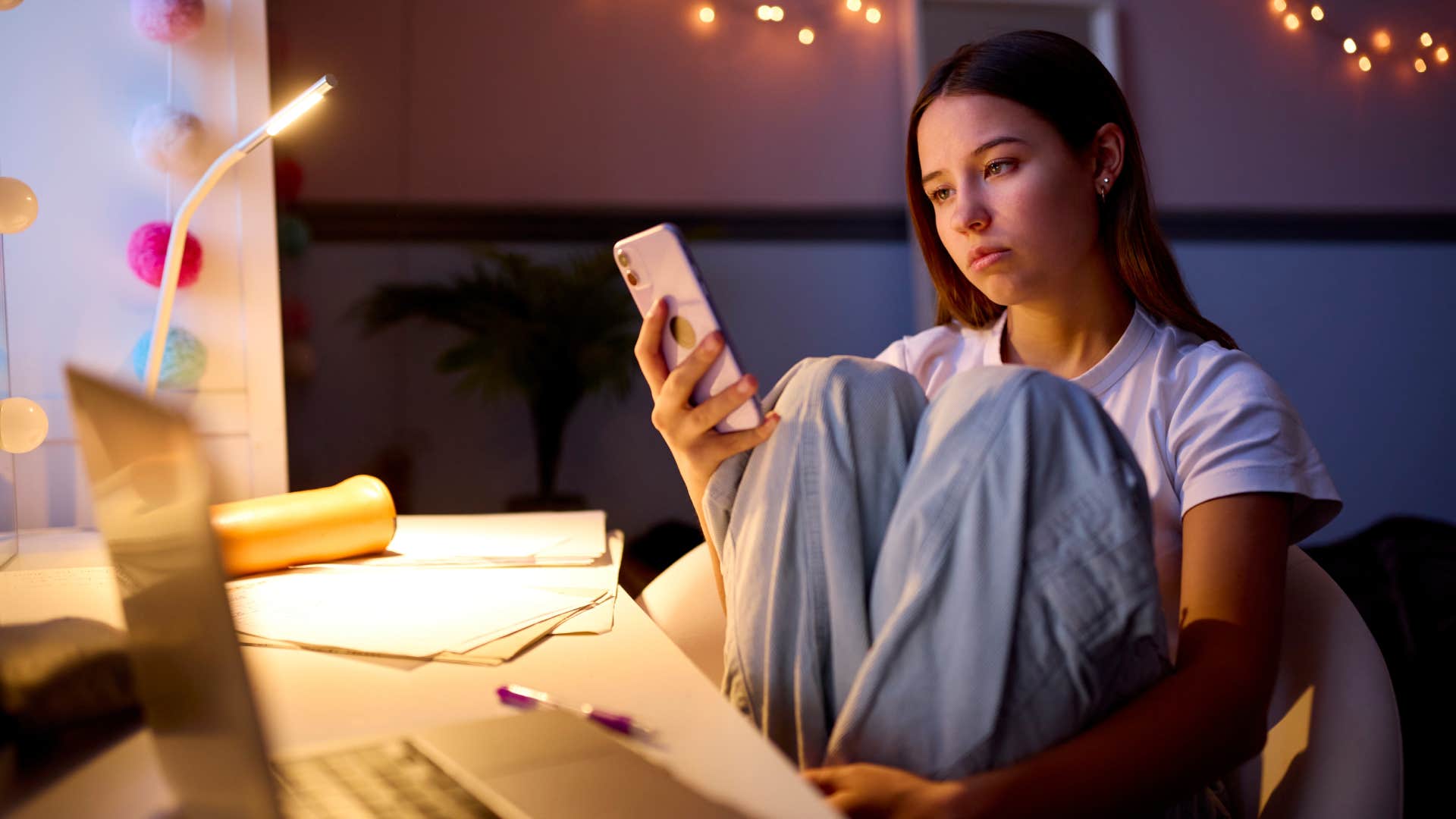 Worried Teenage Girl Sitting At Desk In Bedroom At Home Looking At Mobile Phone At Night