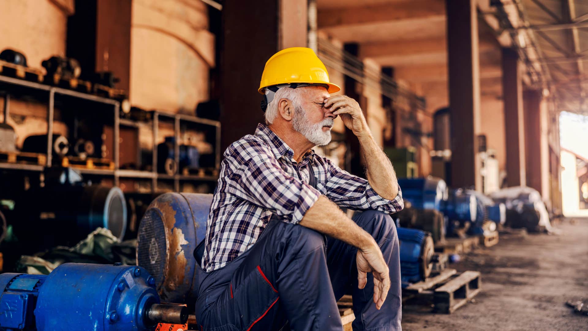 A worried senior factory worker is sitting next to the machines and holding his head