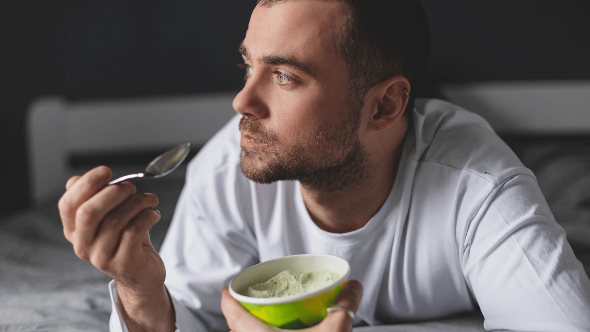 Handsome man lies on his bed while eating a pint of pistachio ice cream