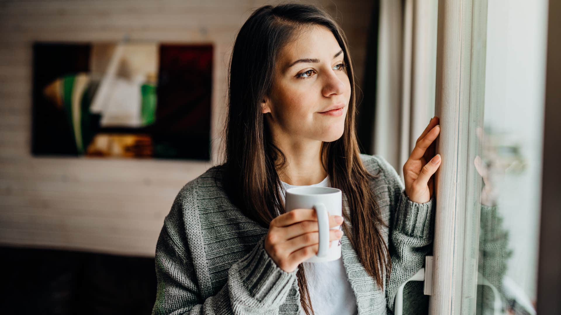 Woman smiling while drinking coffee and looking out a window.