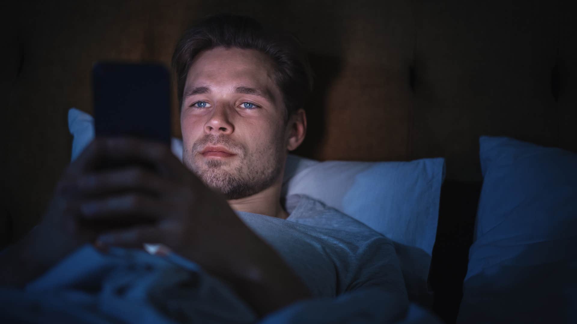 Man scrolling on his phone in a dark bedroom.