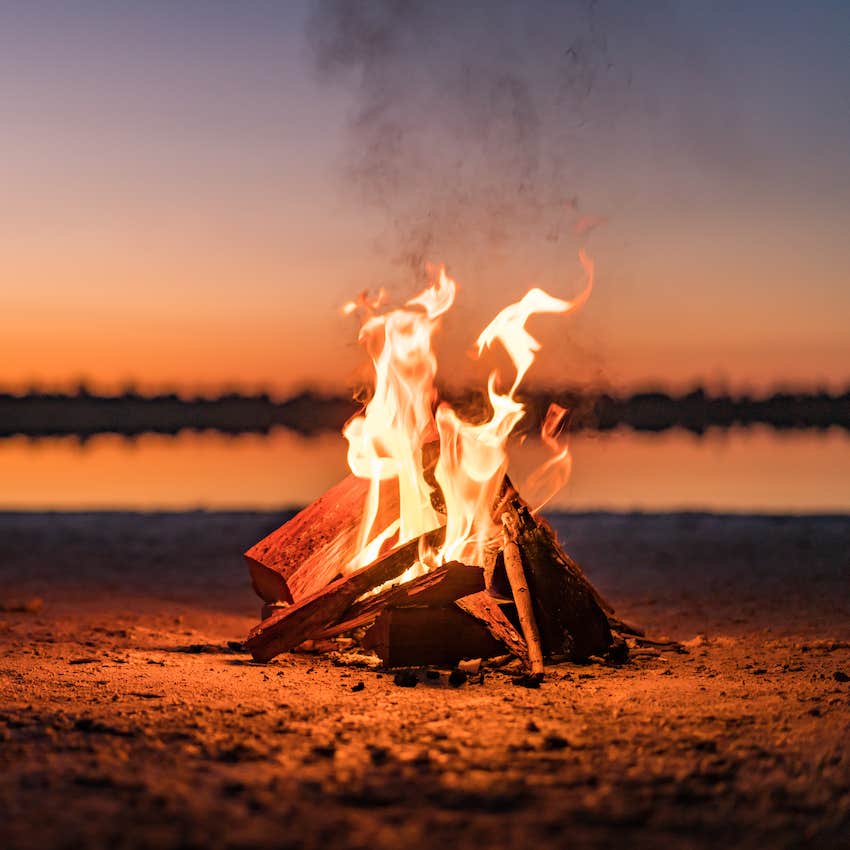 Bonfire burns brightly on sand, water reflected in background