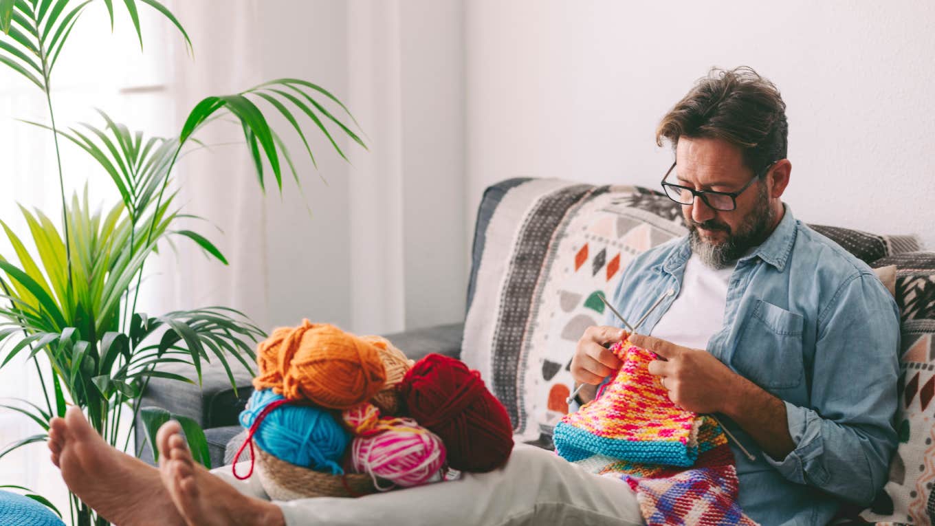 One man at home sitting on the sofa in relaxation indoor leisure activity alone doing knitting work with colorful wool and needles