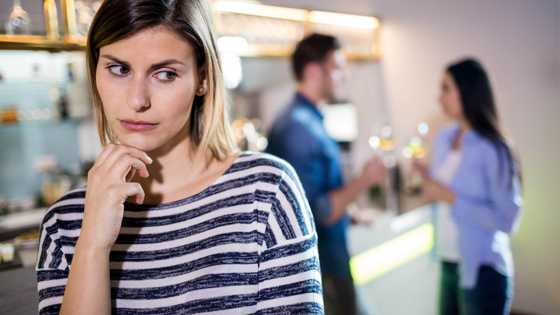 A woman looking jealous as her friend, in the background, sips wine and appears happy in her relationship 