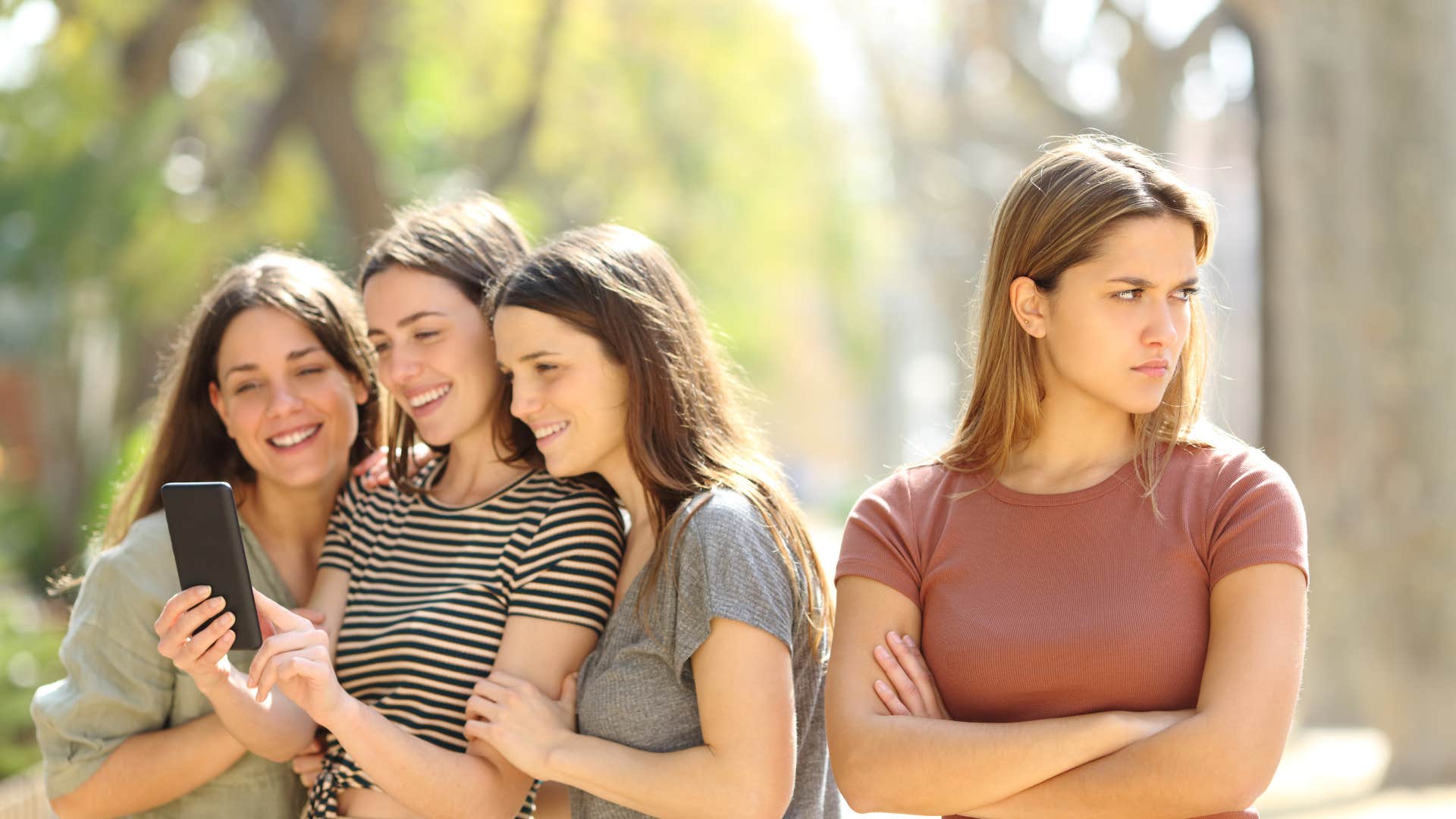 Three girlfriends smiling and looking into the phone camera, while one friend stands aside, seemingly excluded from their plans