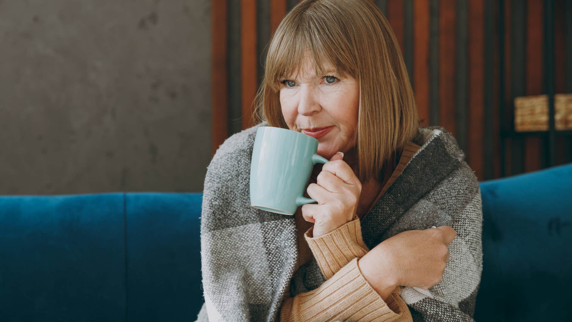 Older woman bundled up sipping from a mug at home