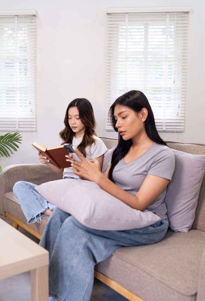 two friends sitting on a couch one reading the other looking at phone