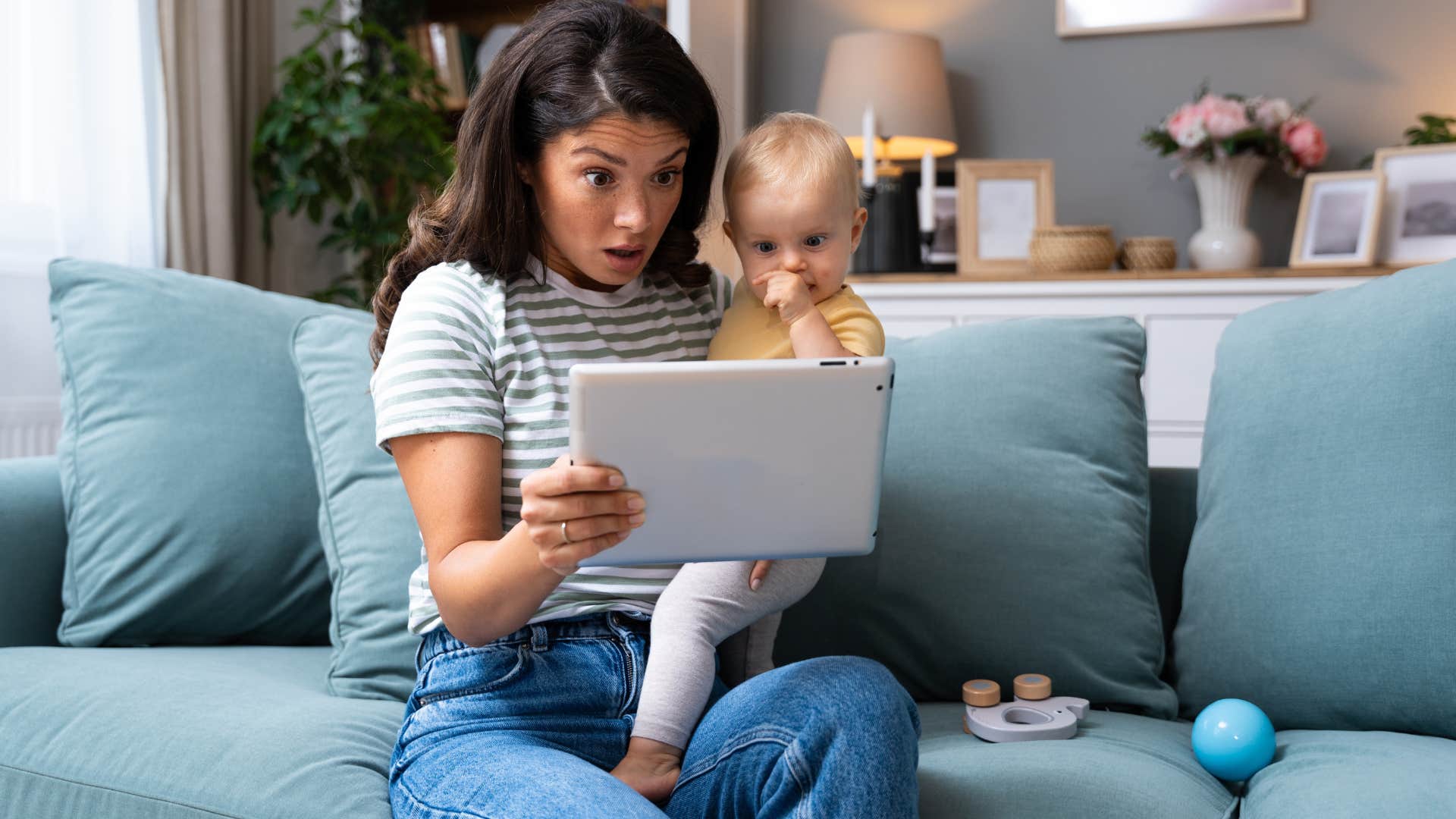 woman holding baby while looking at tablet in shock