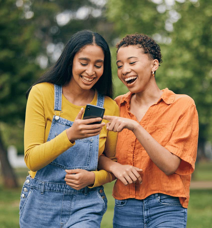 Two happy women look at mobile device while walking in park