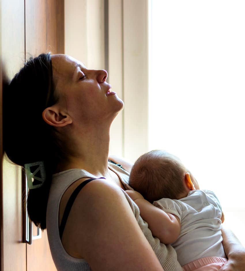 Exhausted woman holding baby leans against a wall