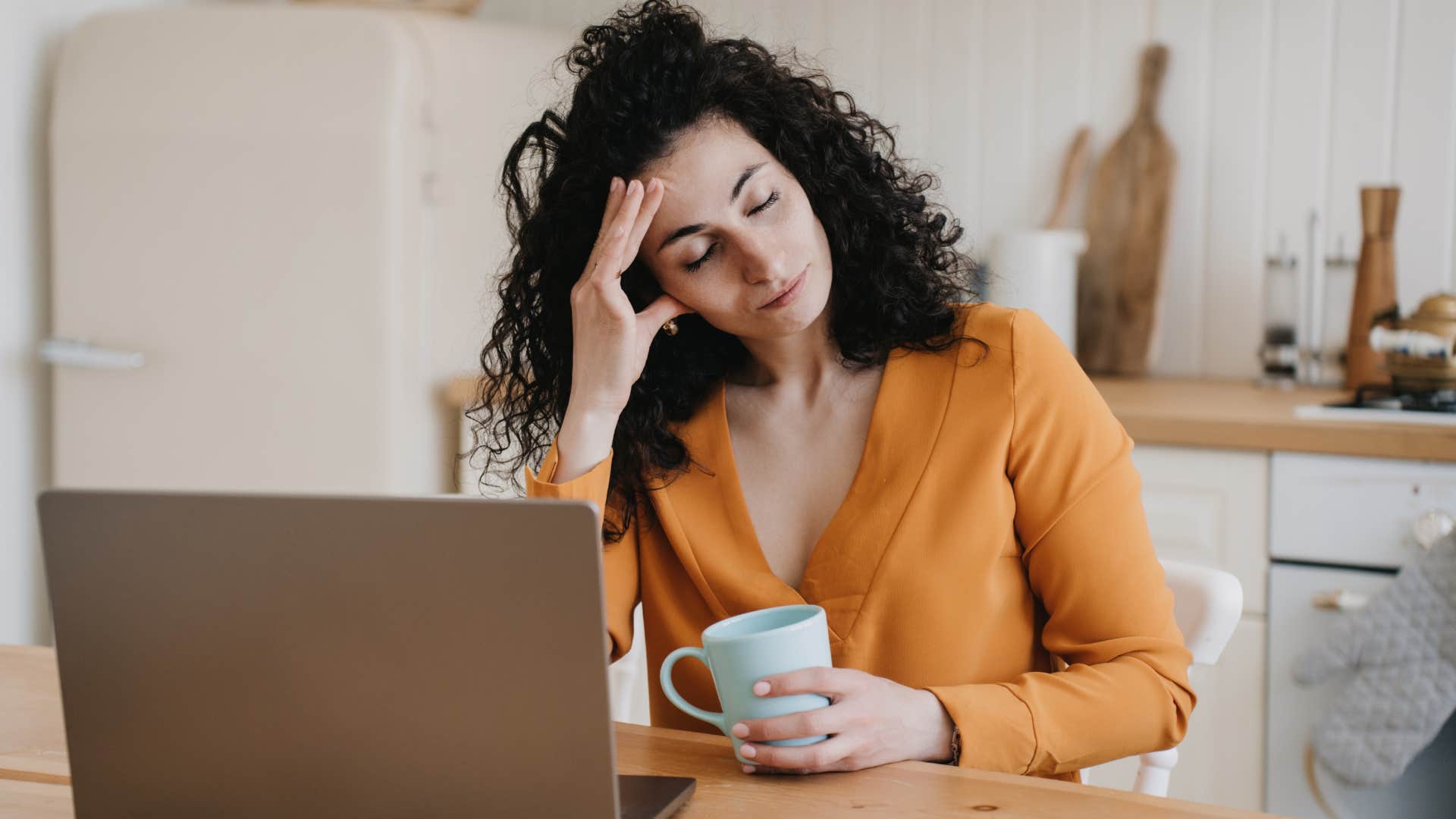 Tired woman yellow with eyes closed sitting at table with laptop holding cup of tea at kitchen