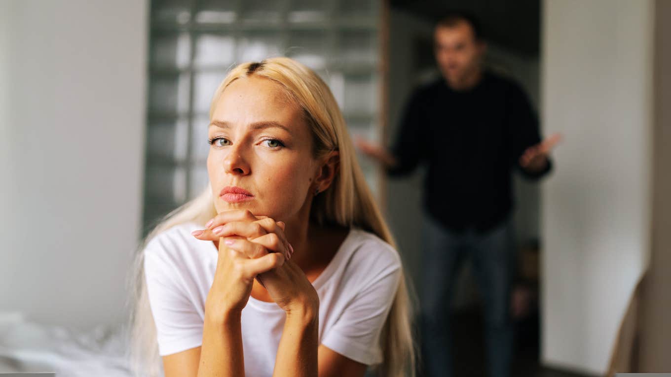 Stoic woman sitting on bed, upset husband trying to control her.