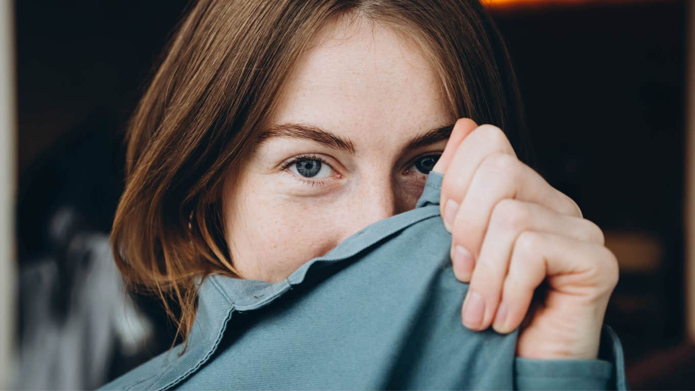 A young woman with blue eyes and freckles on her face smiles and hides her face in a blue shirt.