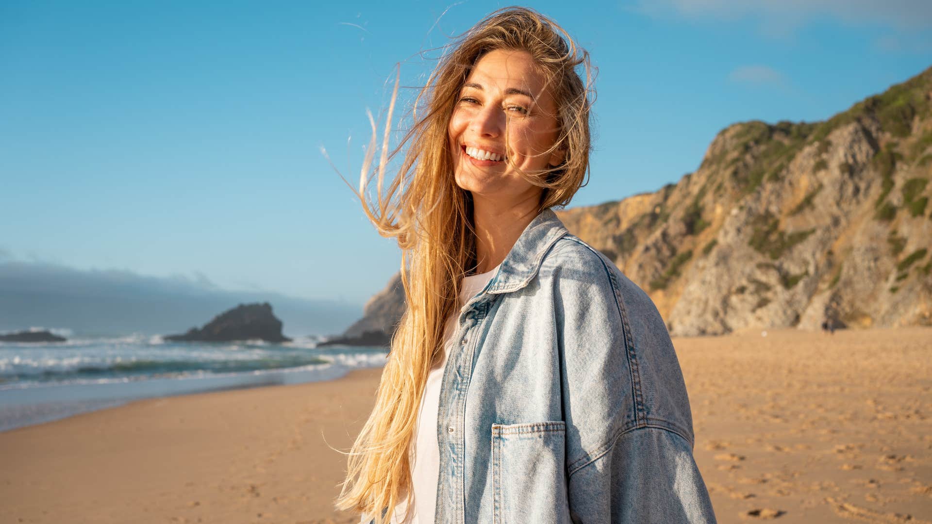 Portrait of smiling woman with long blond hair on sandy beach. 