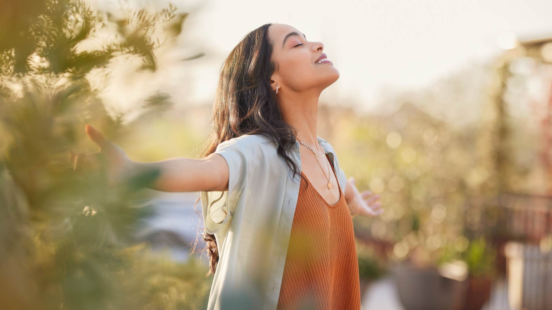 Young latin woman with arms outstretched breathing in fresh air during sunrise at the balcony.