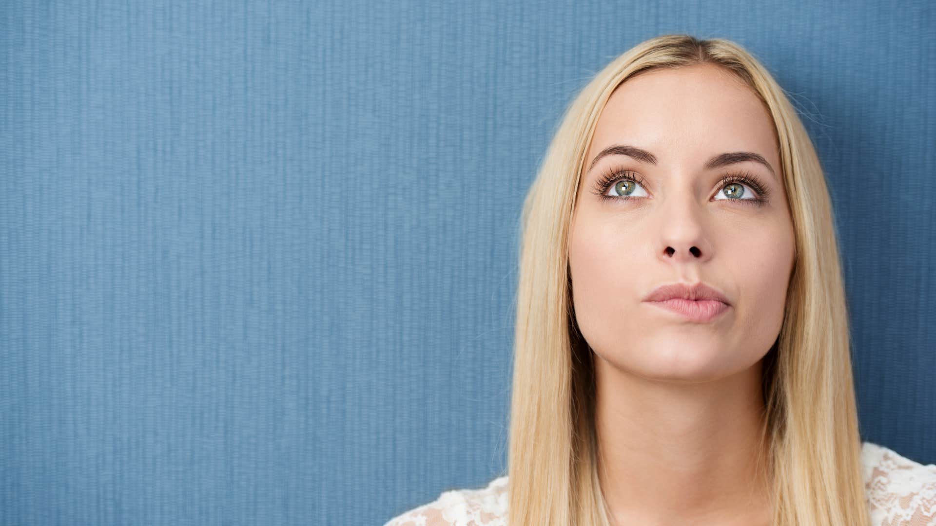 Thoughtful woman with long straight blond hair standing biting her lip as she stares upwards lost in thought