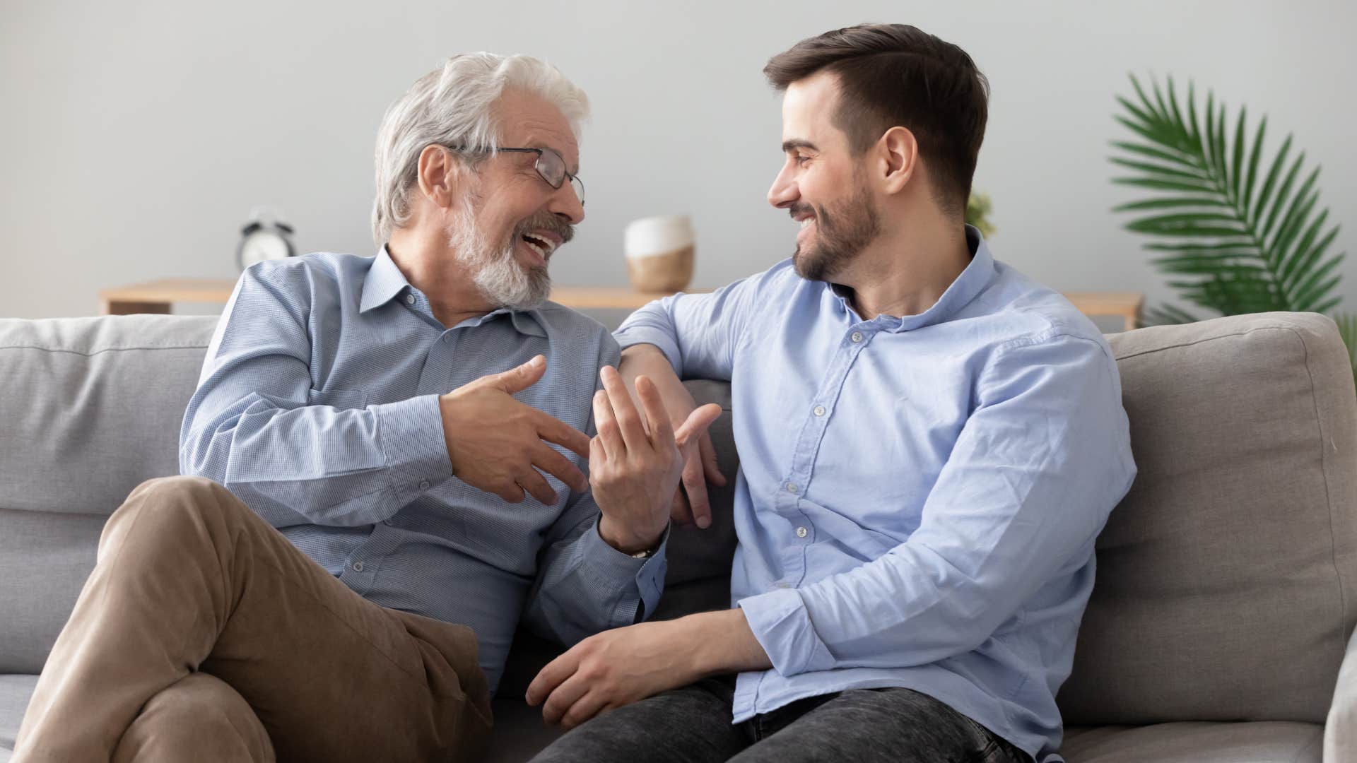 Dad smiling and talking to his adult son on the couch.