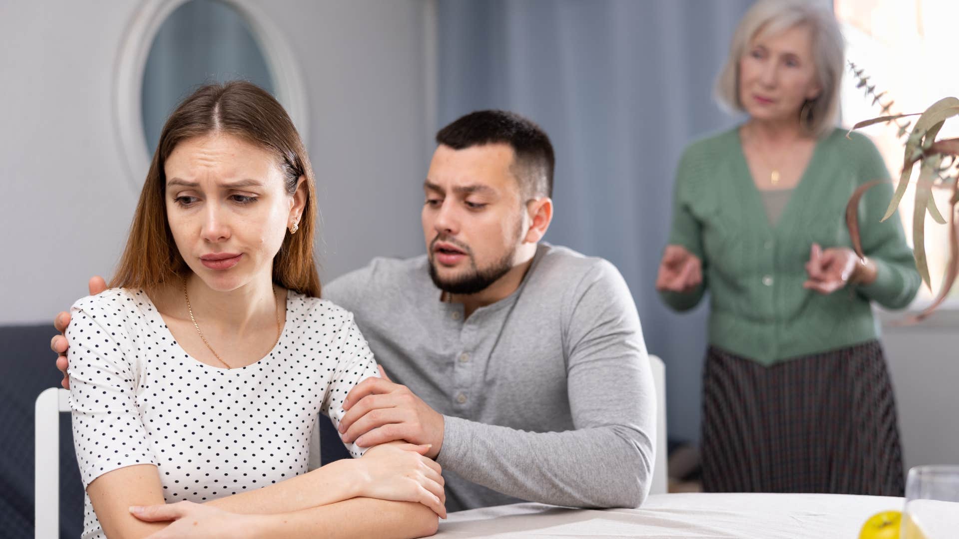 Man comforting his upset wife while his mother argues behind them.