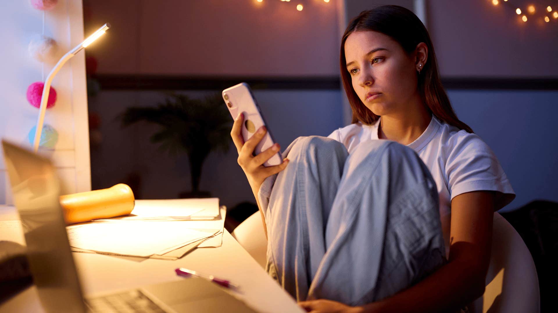 Worried Teenage Girl Sitting At Desk In Bedroom At Home Looking At Mobile Phone At Night