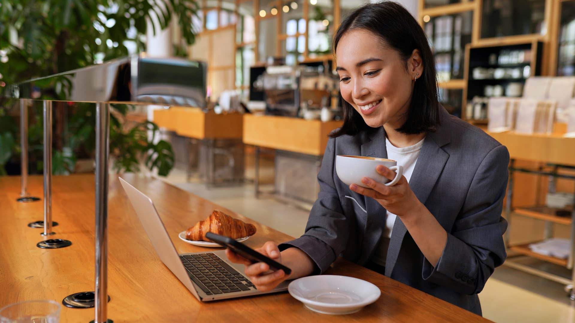 Young Asian business woman wearing suit drinking coffee using smartphone in cafe