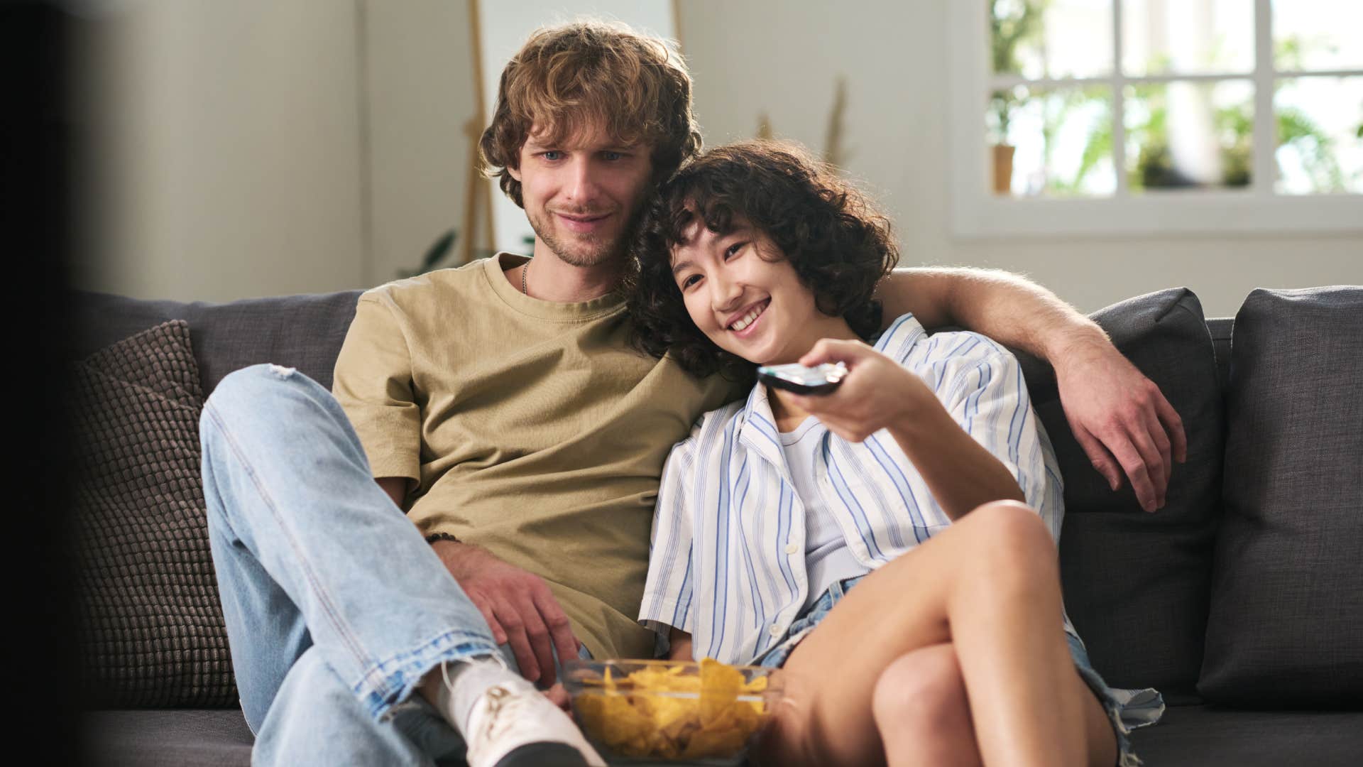 Young woman with remote control and bowl of potato chips choosing amusing tv channel while sitting on couch by her husband