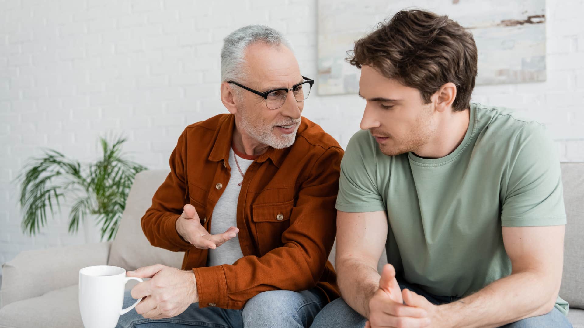 grey haired man holding tea cup and talking to young son on sofa in living room