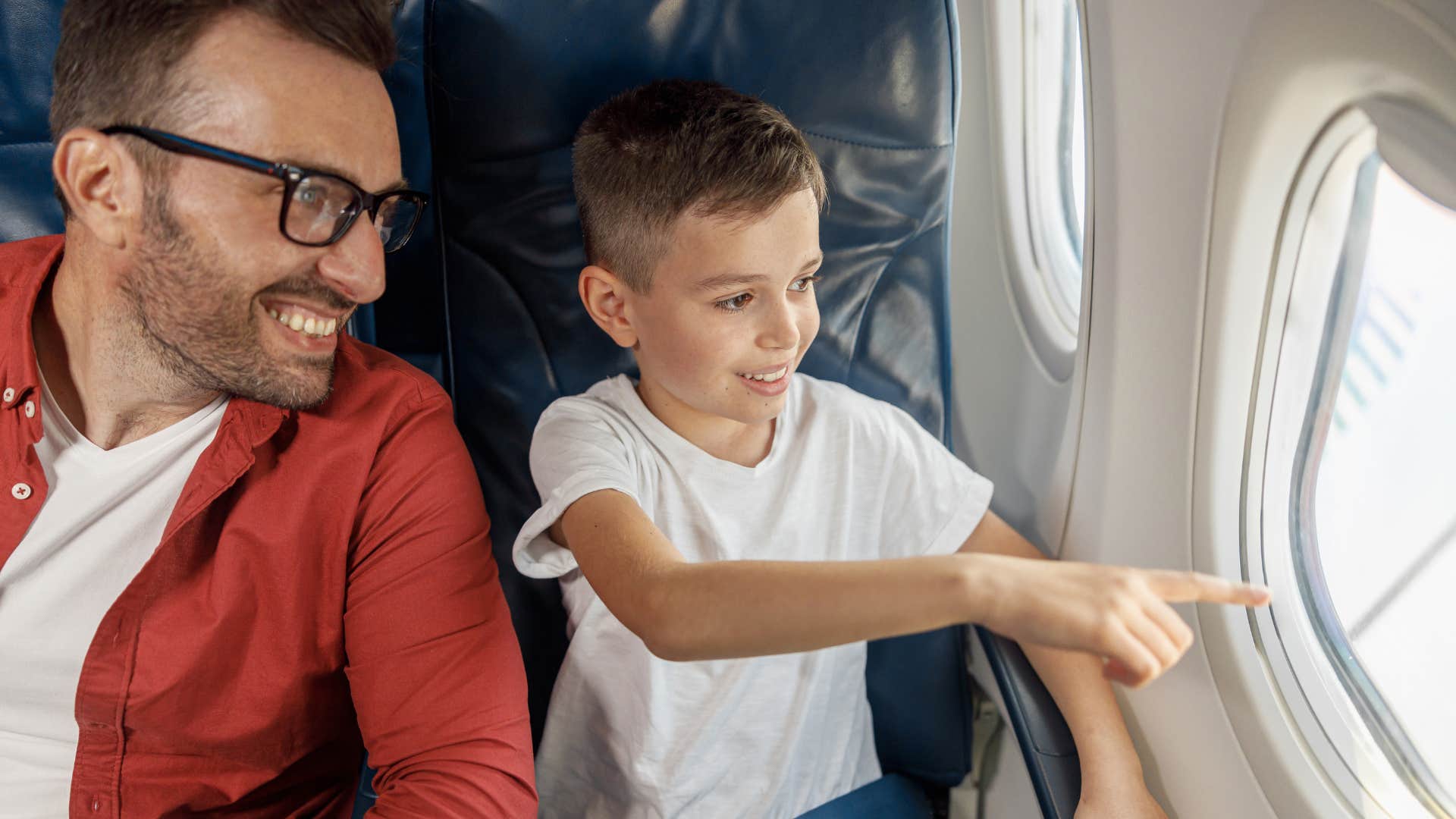 Little boy smiling and pointing out an airplane window next to his dad