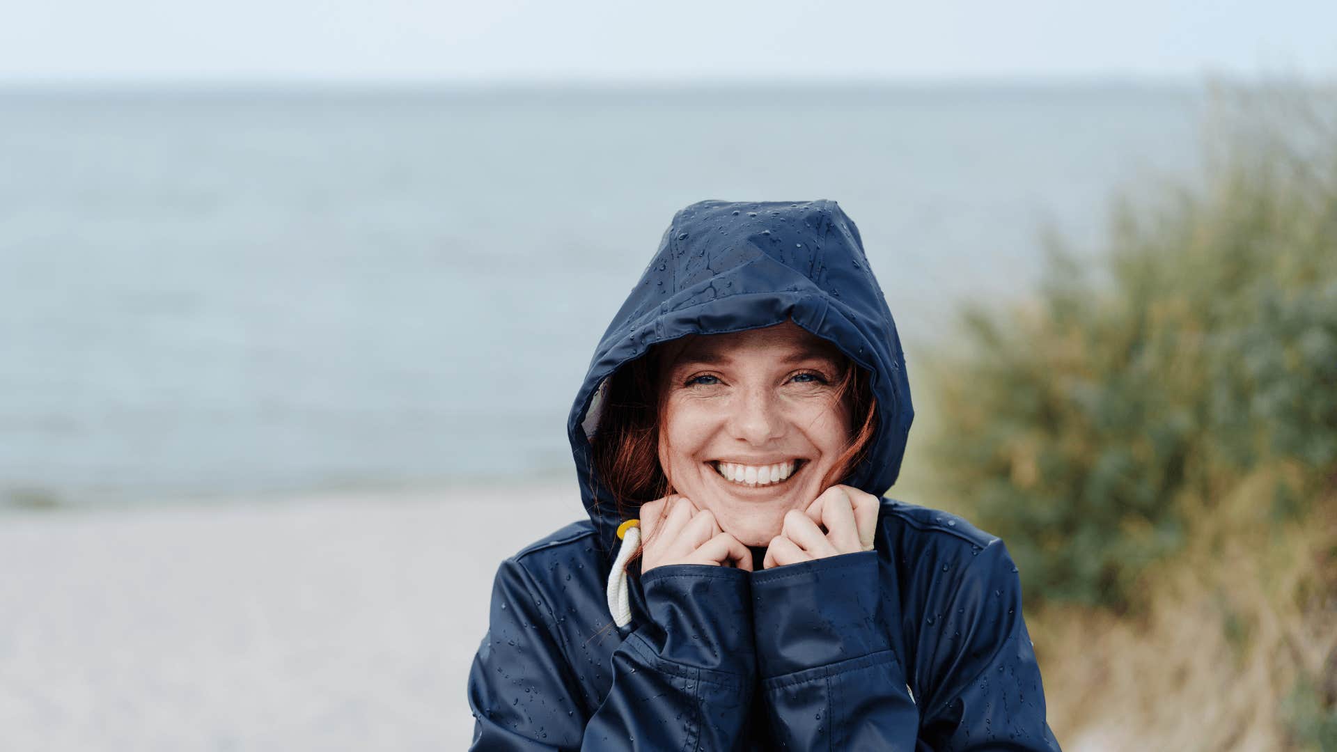 Happy charismatic young woman snuggling into her warm anorak hood with a beaming smile on a cold day at a sandy beach