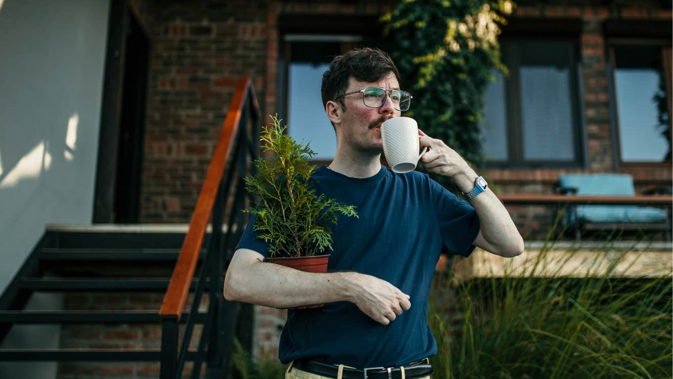 man drinking coffee in front of his house holding a small pine tree