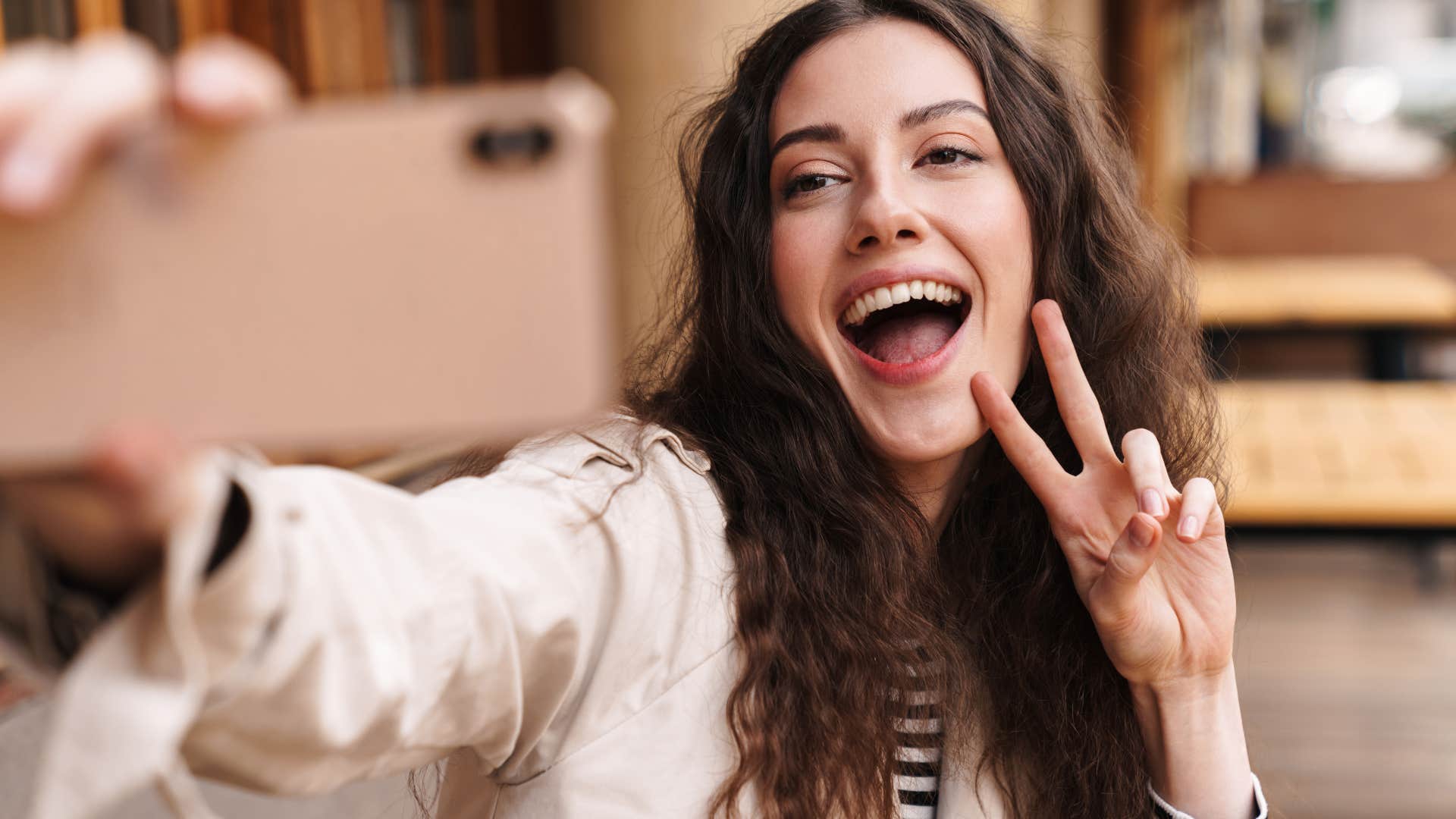 Image of happy brunette woman taking selfie on cellphone and gesturing peace sign in street cafe outdoors