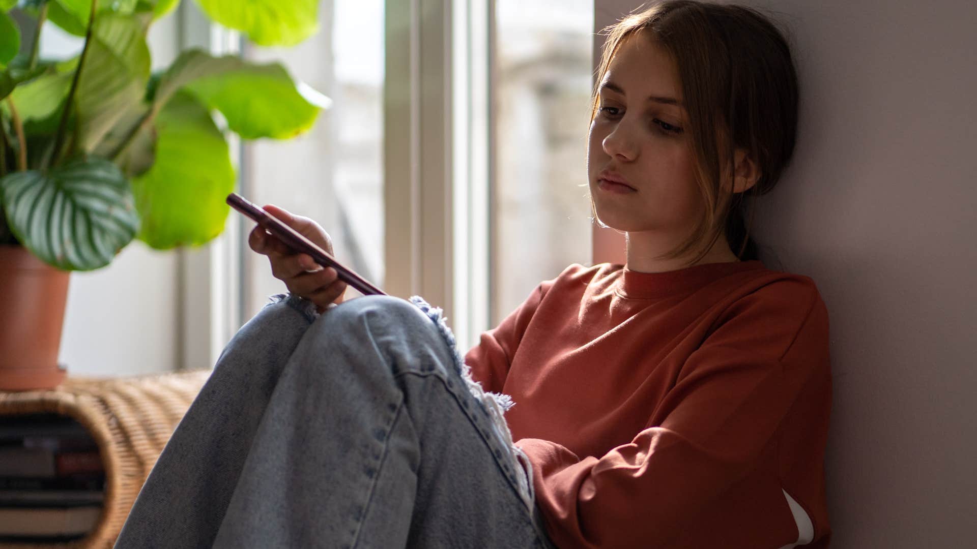 Sad young woman sitting on the floor looking at her phone