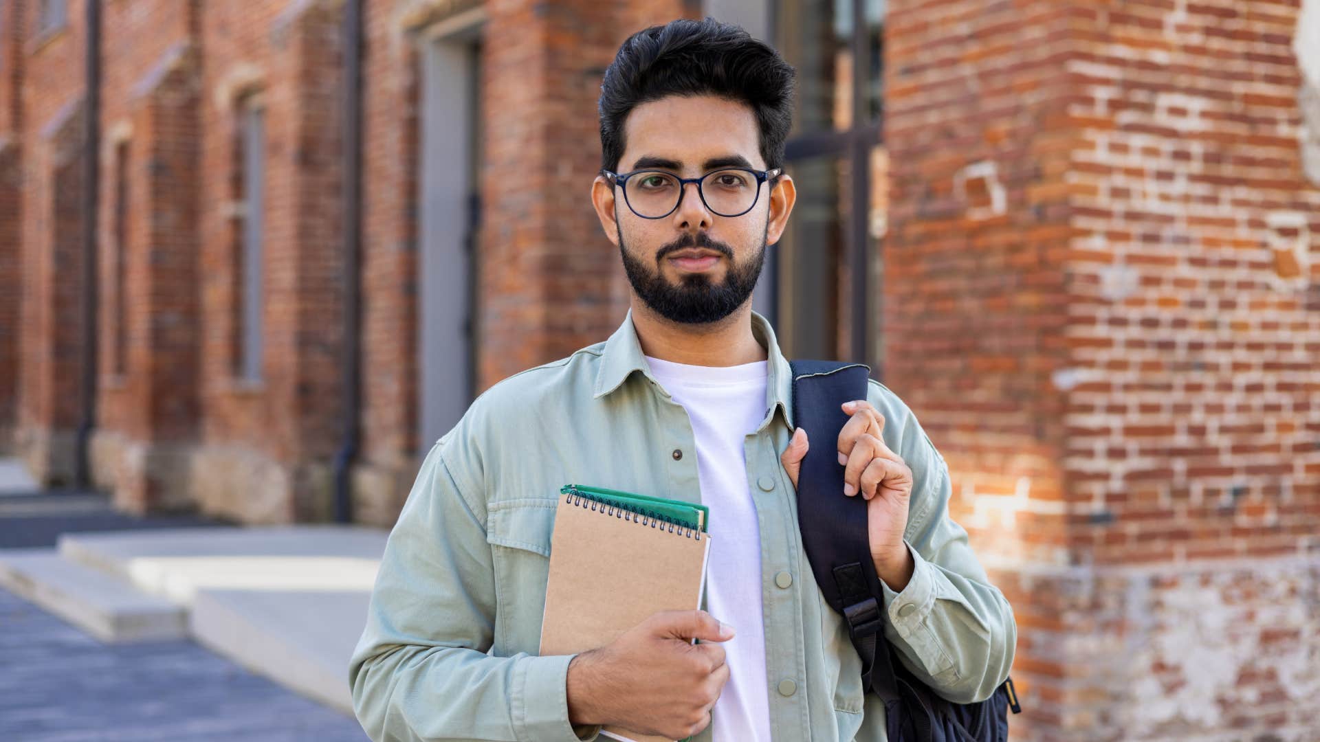 Confident young male Indian student standing outside a university campus with a backpack,