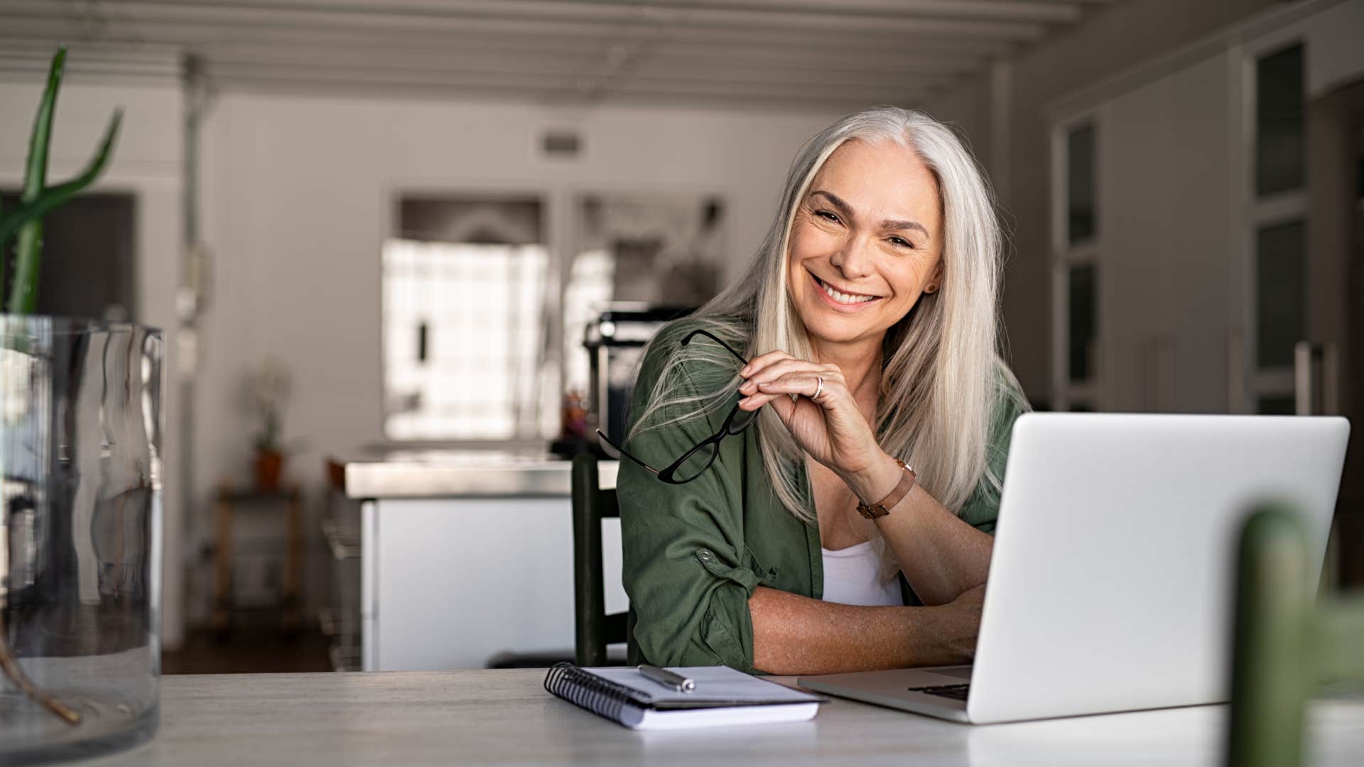 Portrait of happy senior woman holding eyeglasses and looking at camera at home