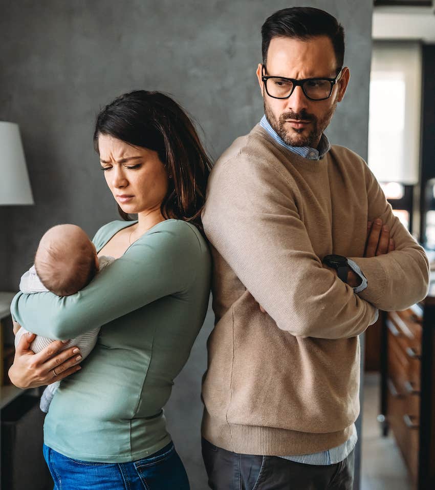 Woman holding infant stand back to back with man who has his arms folded in front of him