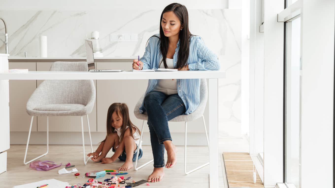 A mom with a smile on her face works while her child is Playing under the desk