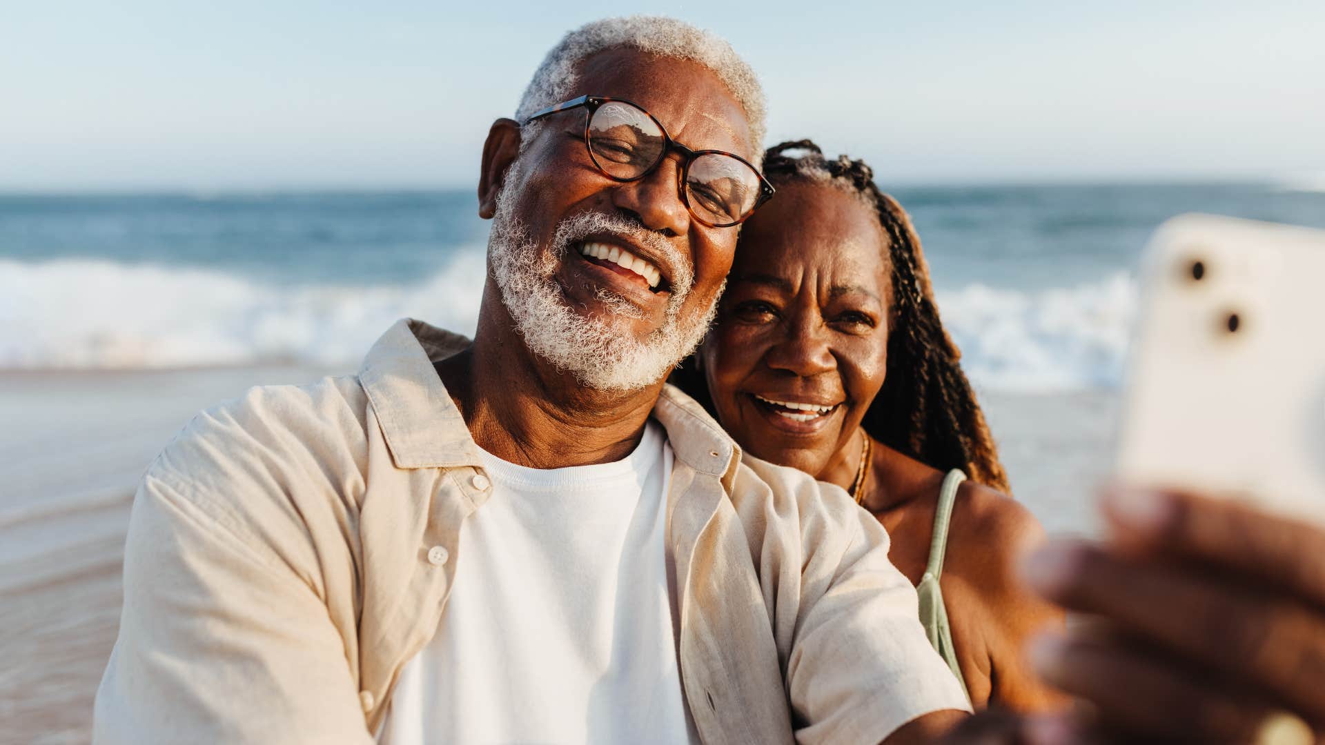 An older couple shares a joyful moment, taking a selfie together with the ocean backdrop during a serene beach walk