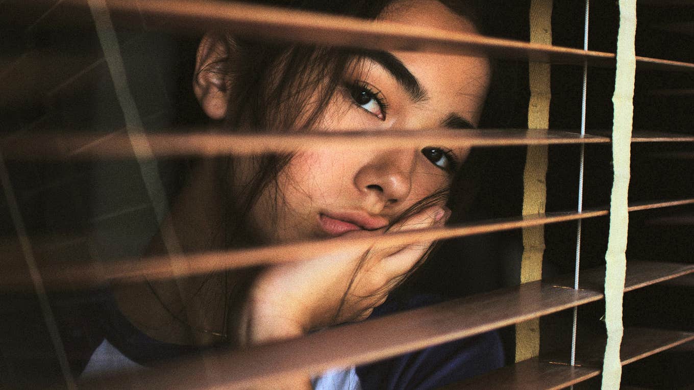 woman looking sadly through window blinds