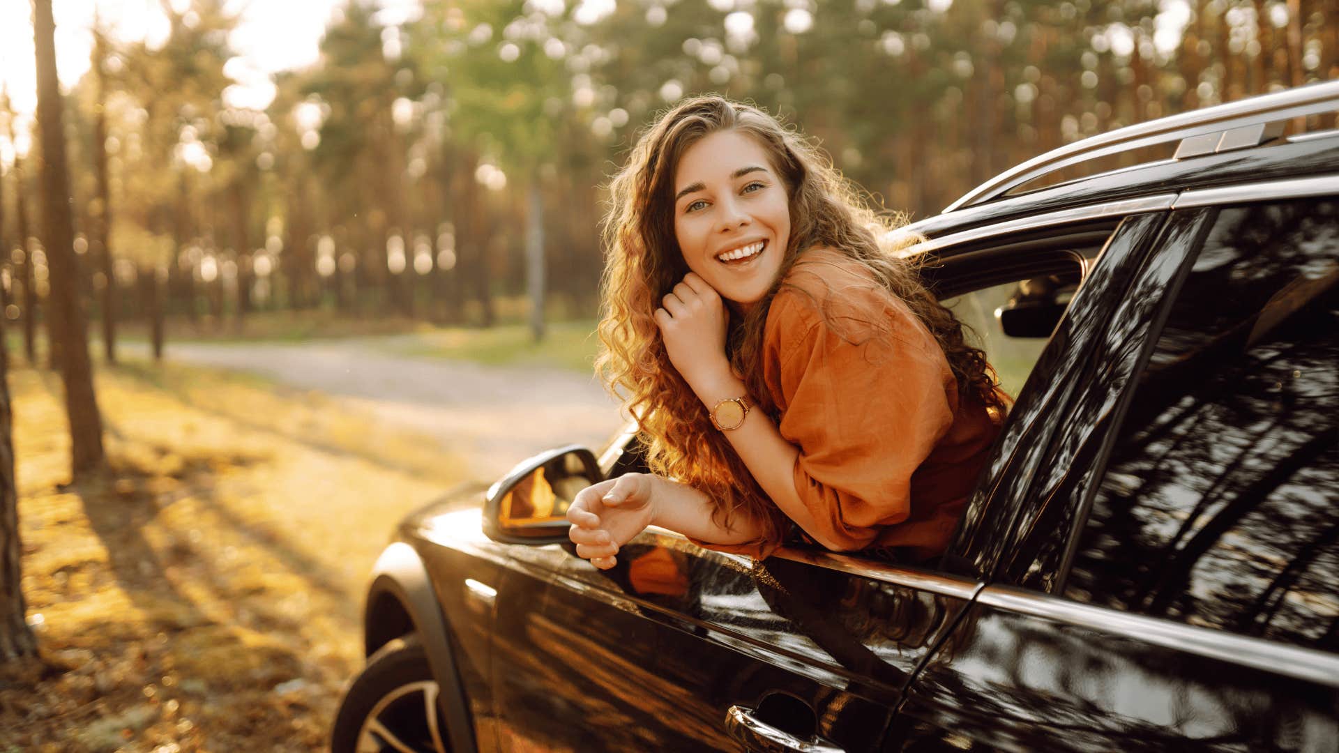 A young woman feels freedom leaning out of a car window in a sunny forest. 