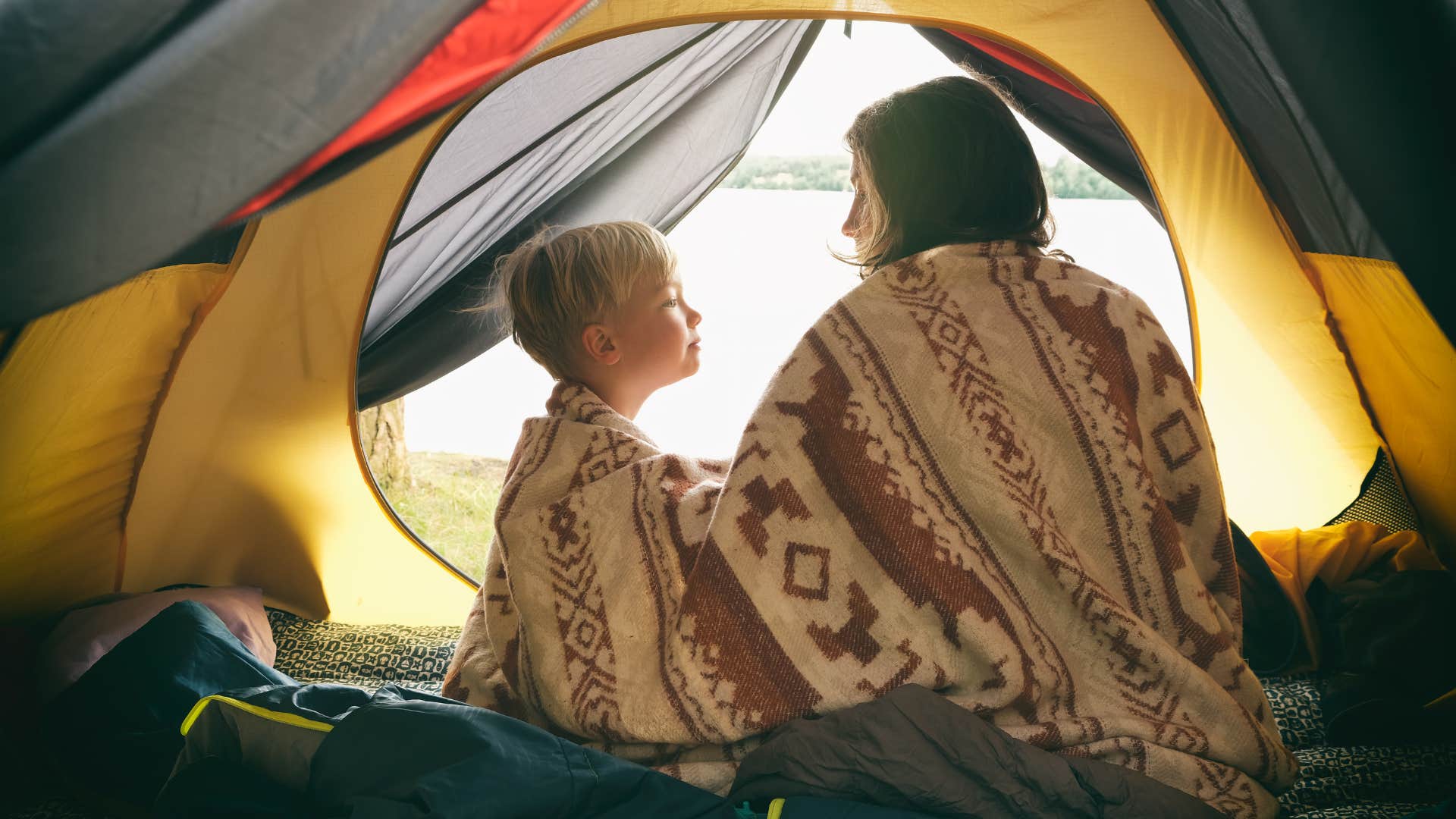 Mother and son sitting in camping tent, wrapped in wool blanket and admiring sunrise on the river