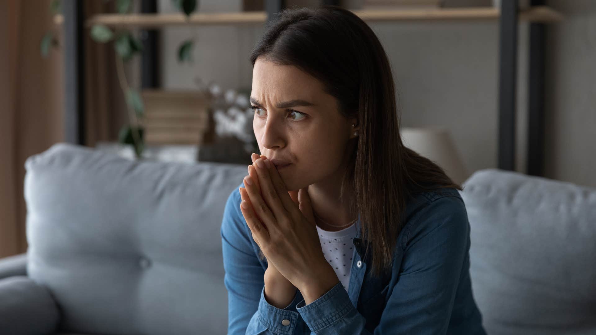 Worried young woman sitting on couch with folded hands