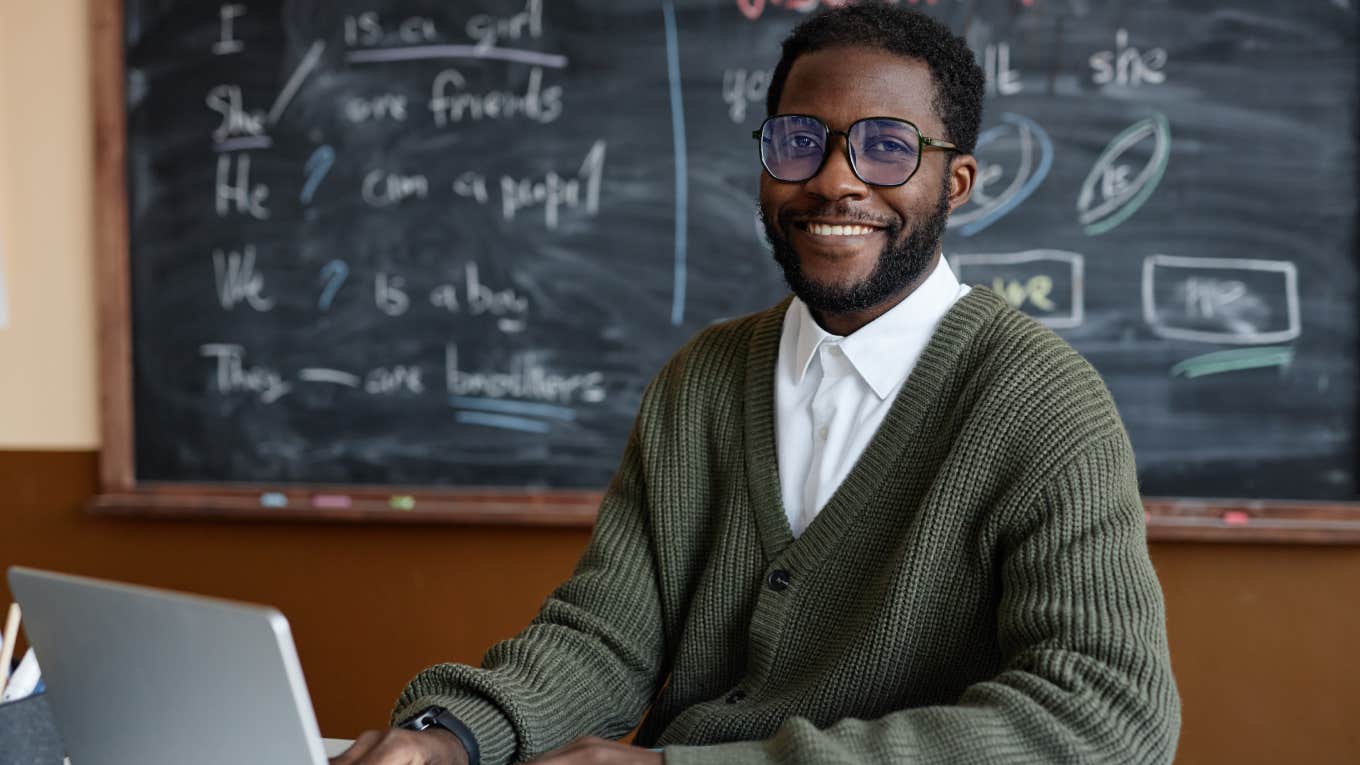 portrait of young African American male teacher of English wearing eyeglasses sitting at desk in classroom smiling at camera while using laptop