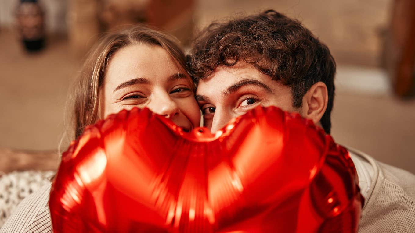 Young couple in love holding a heart-shaped balloon, hiding behind it while sitting on the sofa