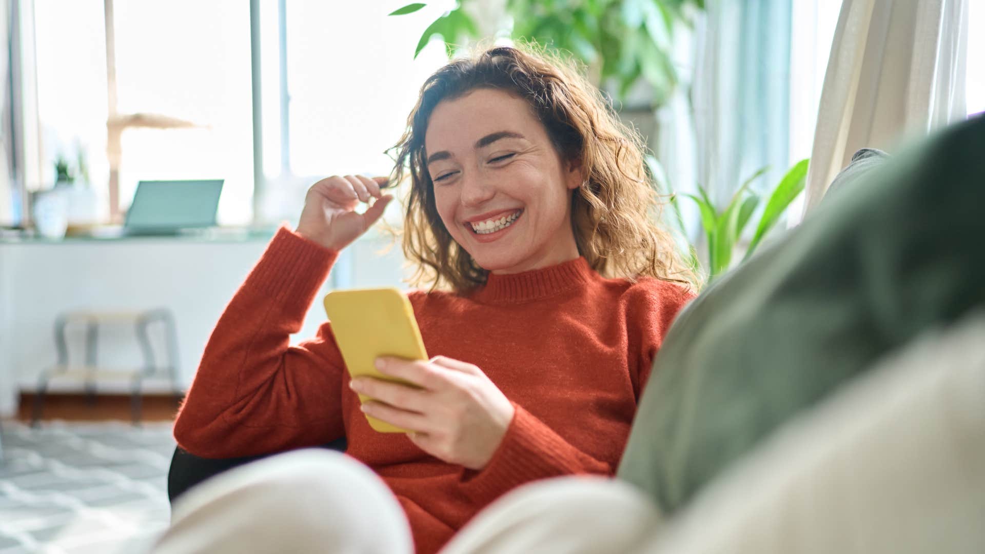 Happy relaxed young woman sitting on couch using cell phone